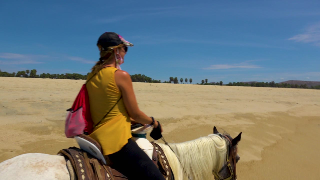 At the start of the San Jose Del Cabo clip, a woman in a yellow top rides a white horse across open pale sand under a clear blue sky.