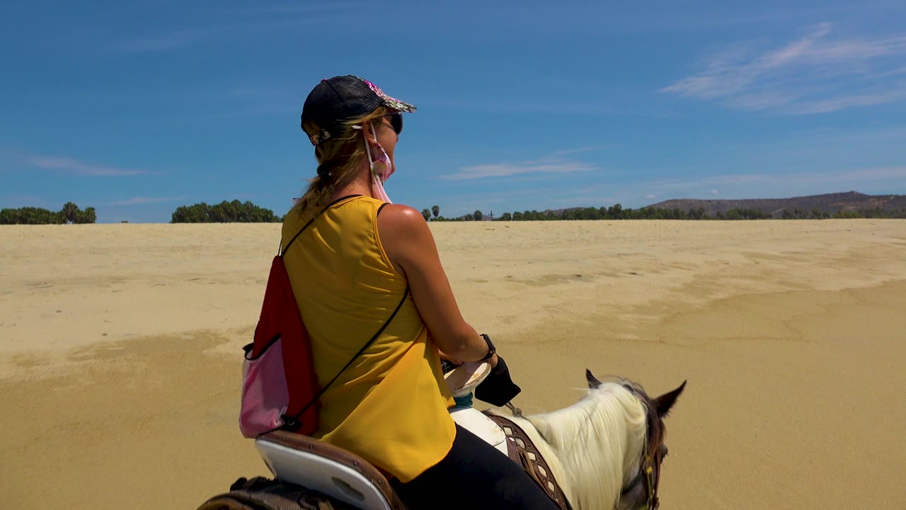 A rider in a yellow top is seen from behind on horseback at San Jose Del Cabo, Mexico, facing broad pale sand under a blue sky.
