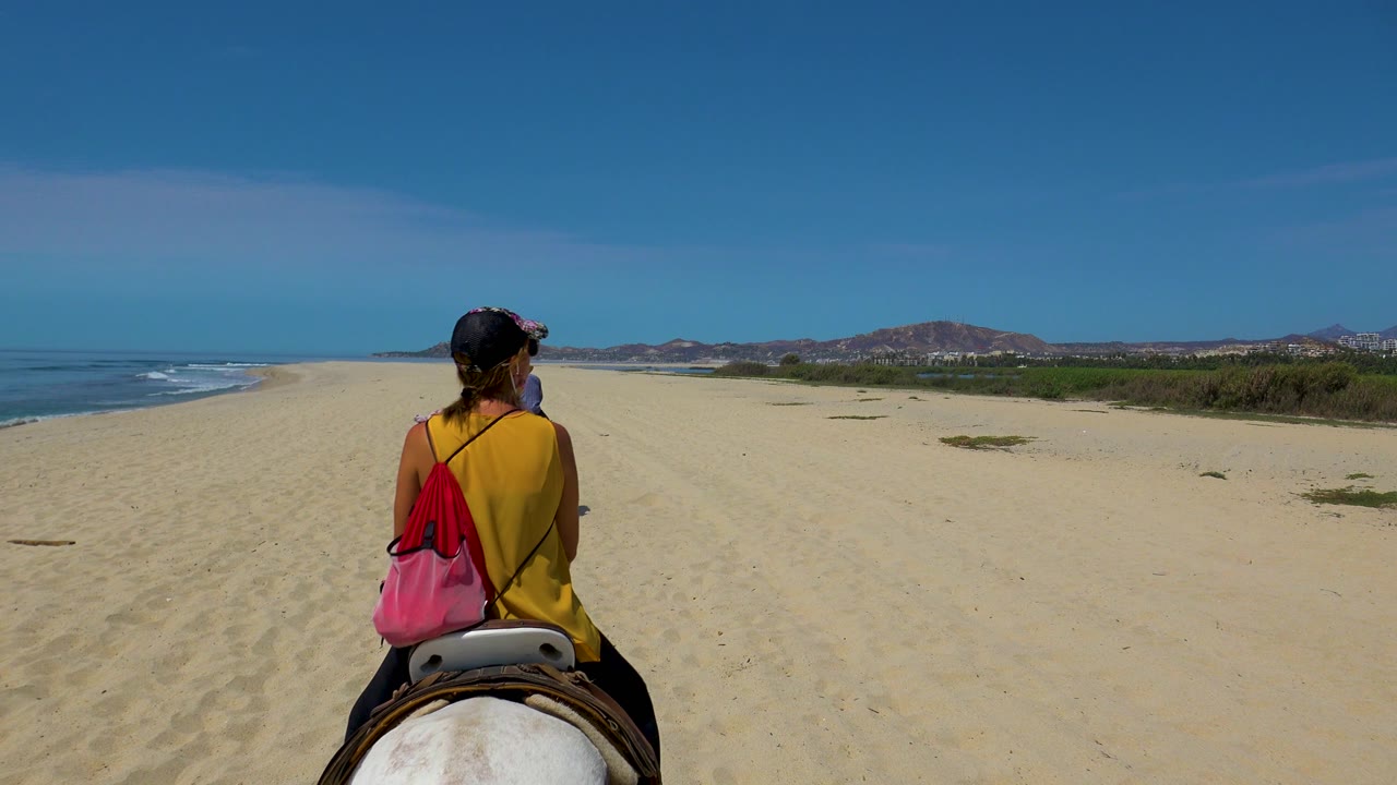 A rear view of the rider and horse on the San Jose del Cabo beach shows more open sand ahead, the ocean breaking along the left edge, and low hills across the far right horizon.
