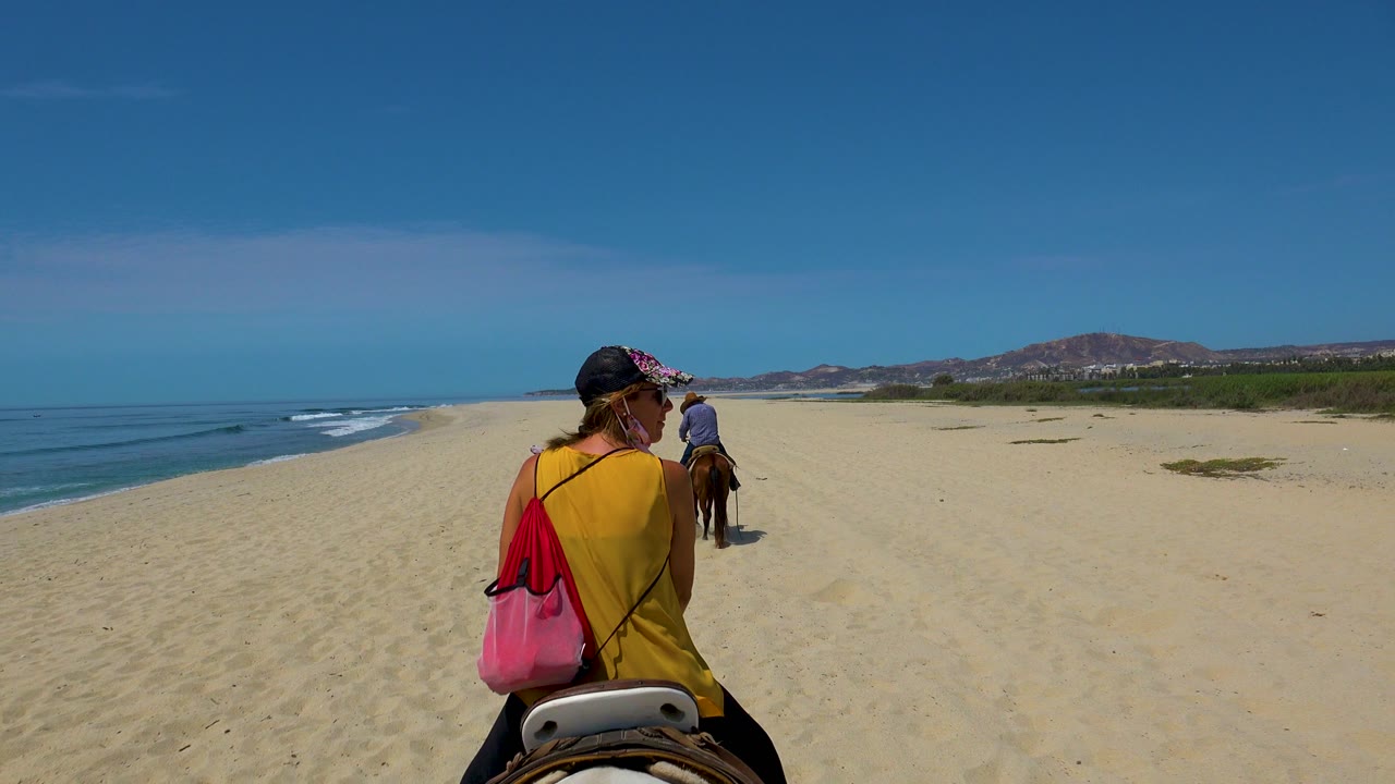 In San Jose del Cabo, a foreground rider in a yellow top follows another rider across pale sand with blue ocean surf on the left and low hills ahead.