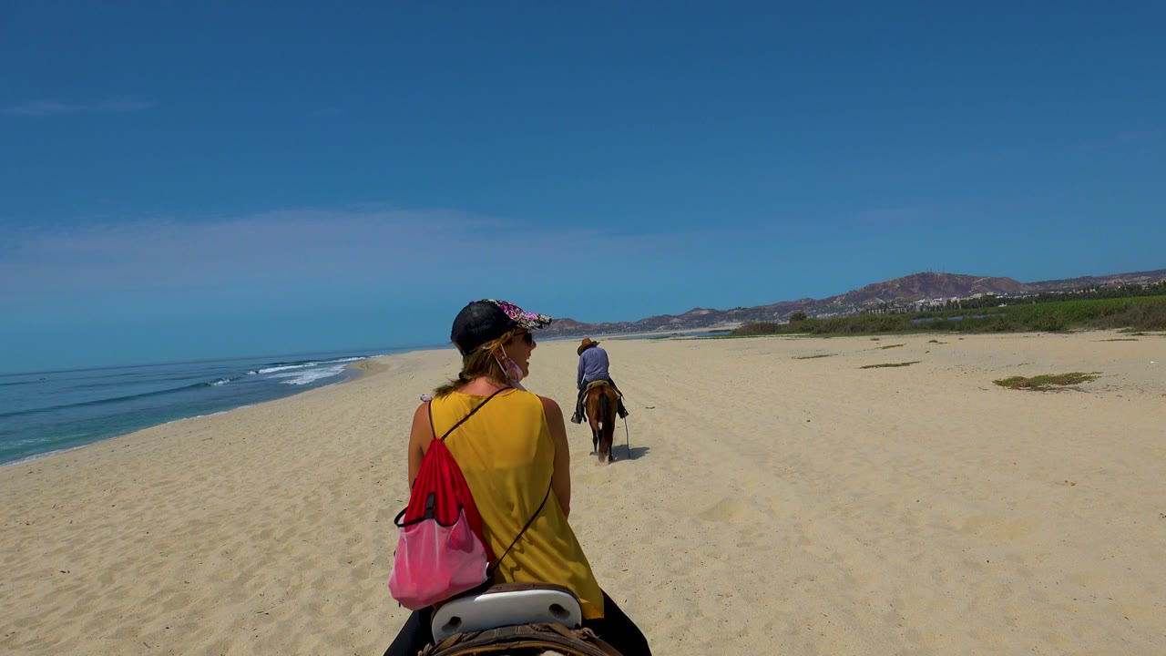 A rider in a yellow top remains in the foreground while another rider moves farther ahead on the sandy San Jose del Cabo beach, with the ocean edge visible at left.