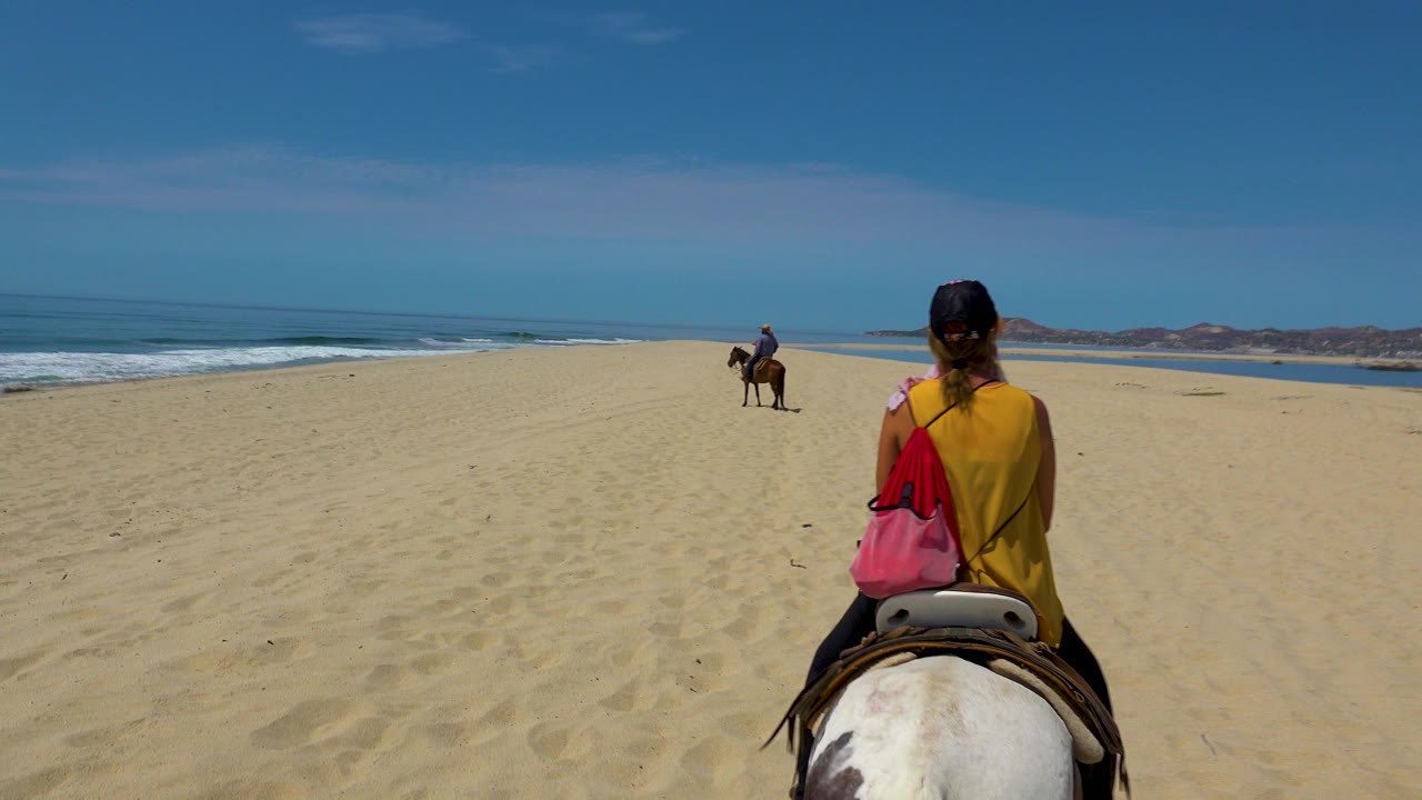 Rear view of a woman riding a white and brown horse on a wide San Jose Del Cabo beach, with the ocean on the left and a distant rider ahead.