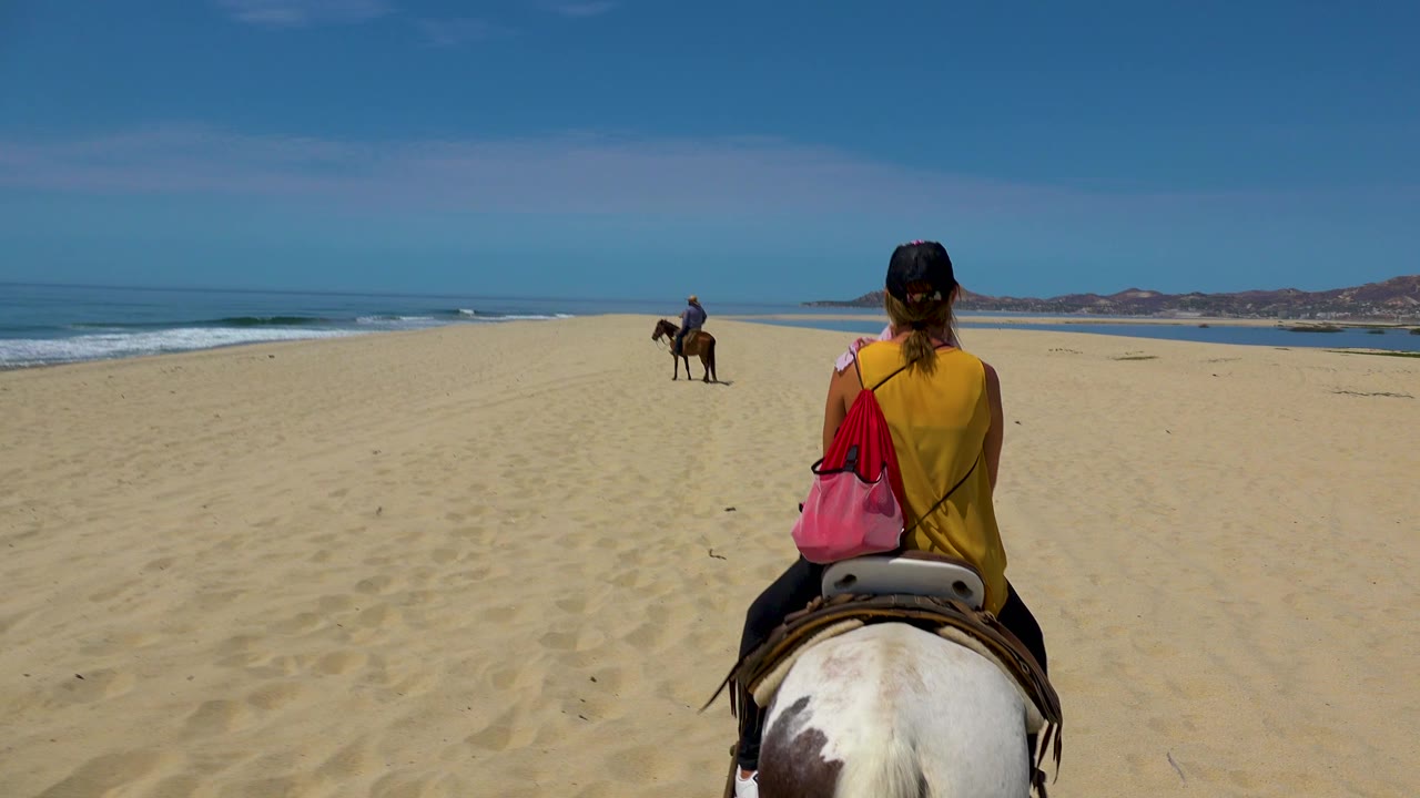 The foreground rider sits on a white and brown horse on the open San Jose Del Cabo sand, with a second rider ahead near the waterline and hills beyond.