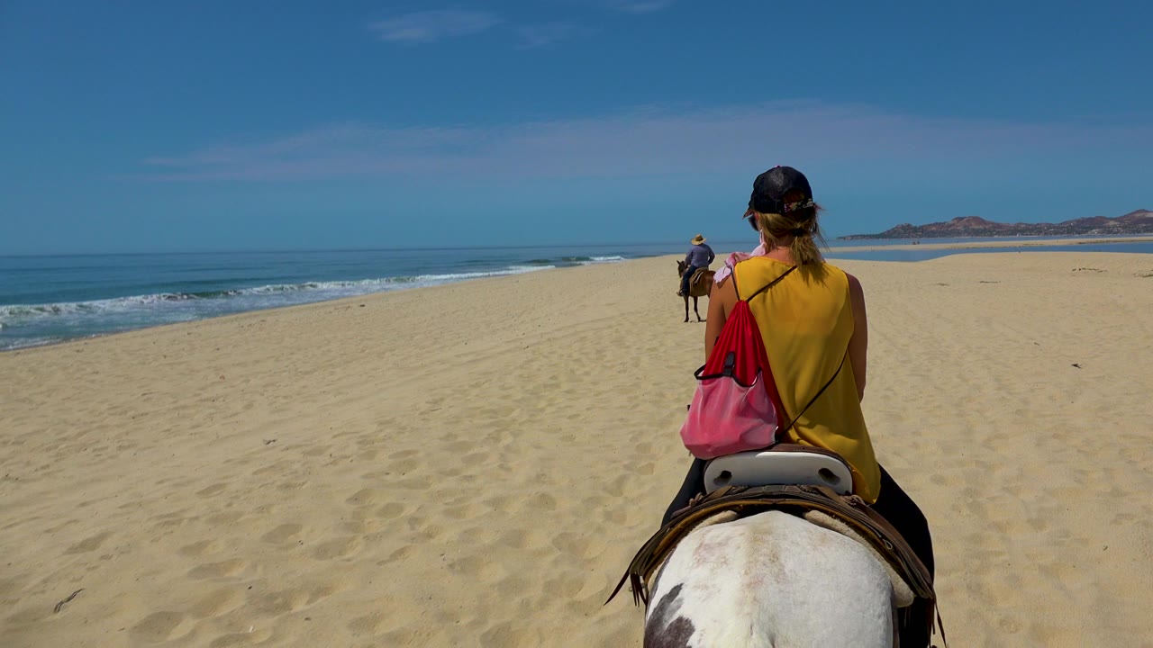 A woman in a yellow top rides a white horse along a wide San Jose Del Cabo beach, with another rider ahead and surf rolling on the left.