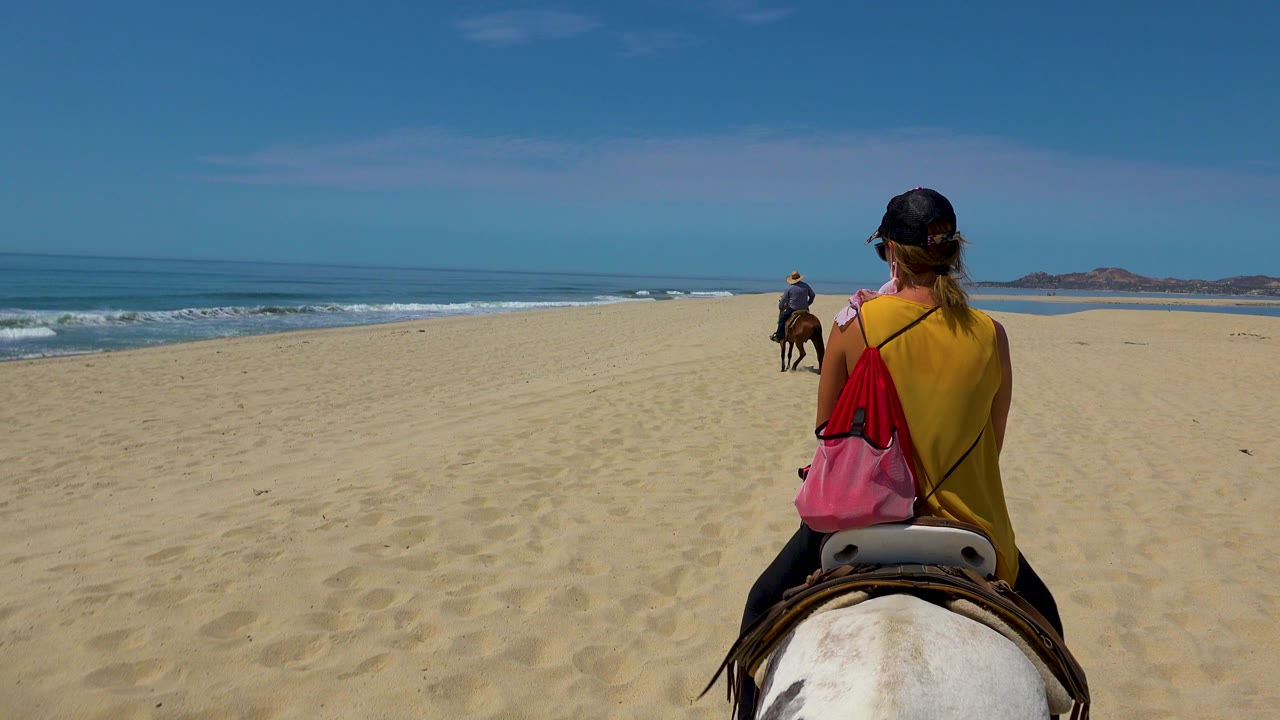 The woman on horseback is framed on the right side, with the ocean and waves to the left and another rider moving ahead across the San Jose Del Cabo sand.