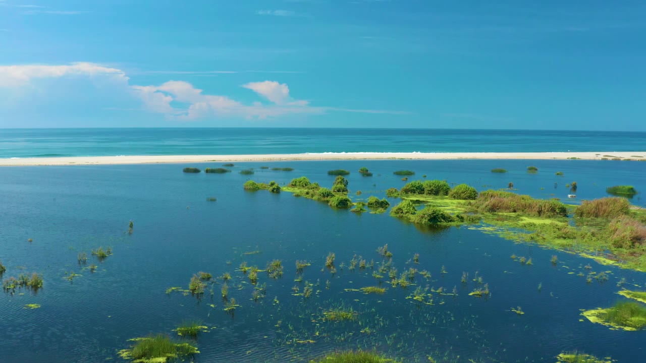 At 0:03, the aerial composition in San Jose del Cabo shows more open blue water in the foreground, green vegetation grouped to the right, a pale sandbar behind it, and no visible people.