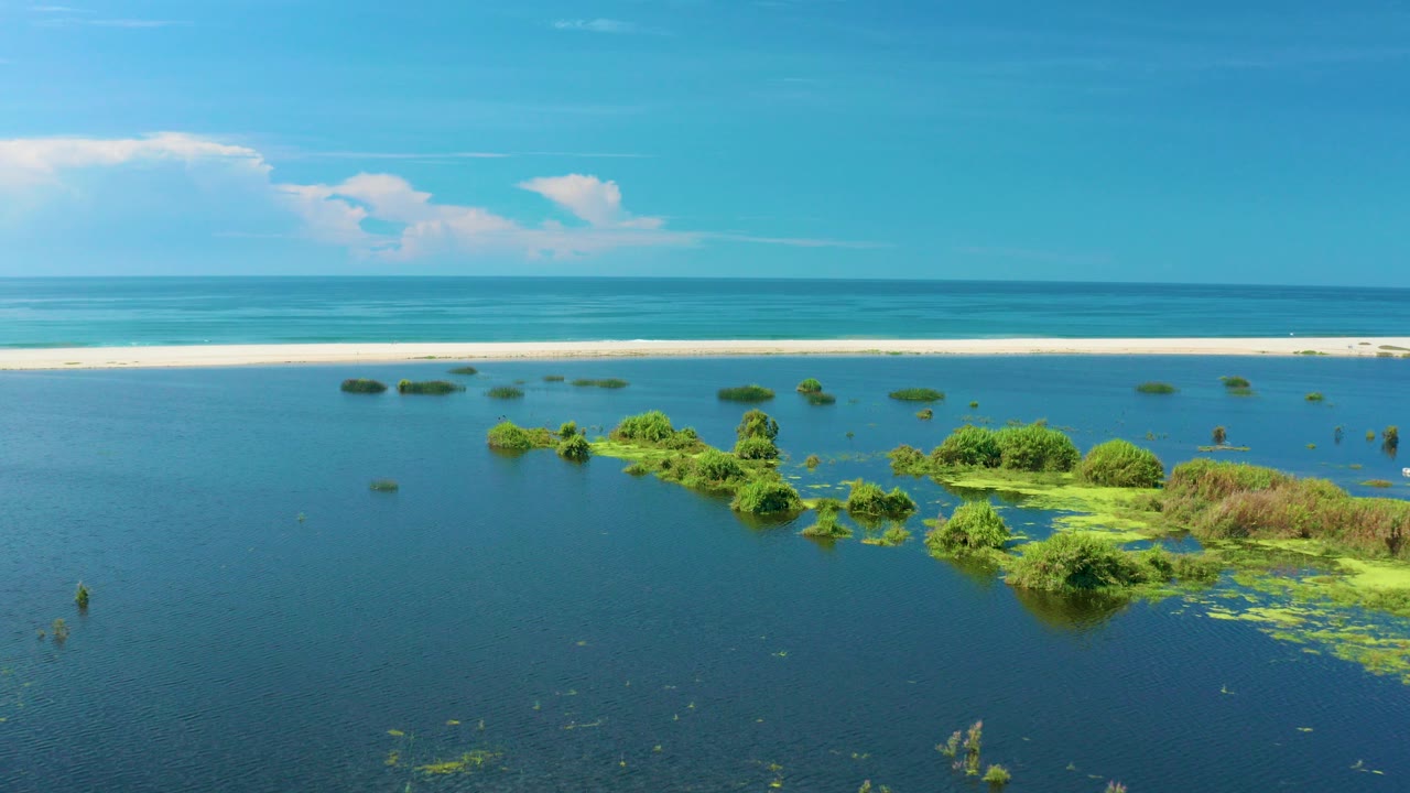 Aerial view in San Jose Del Cabo, Mexico, with blue lagoon water around green vegetated islands, a pale sandbar, distant surf, bright sky, and no visible people.