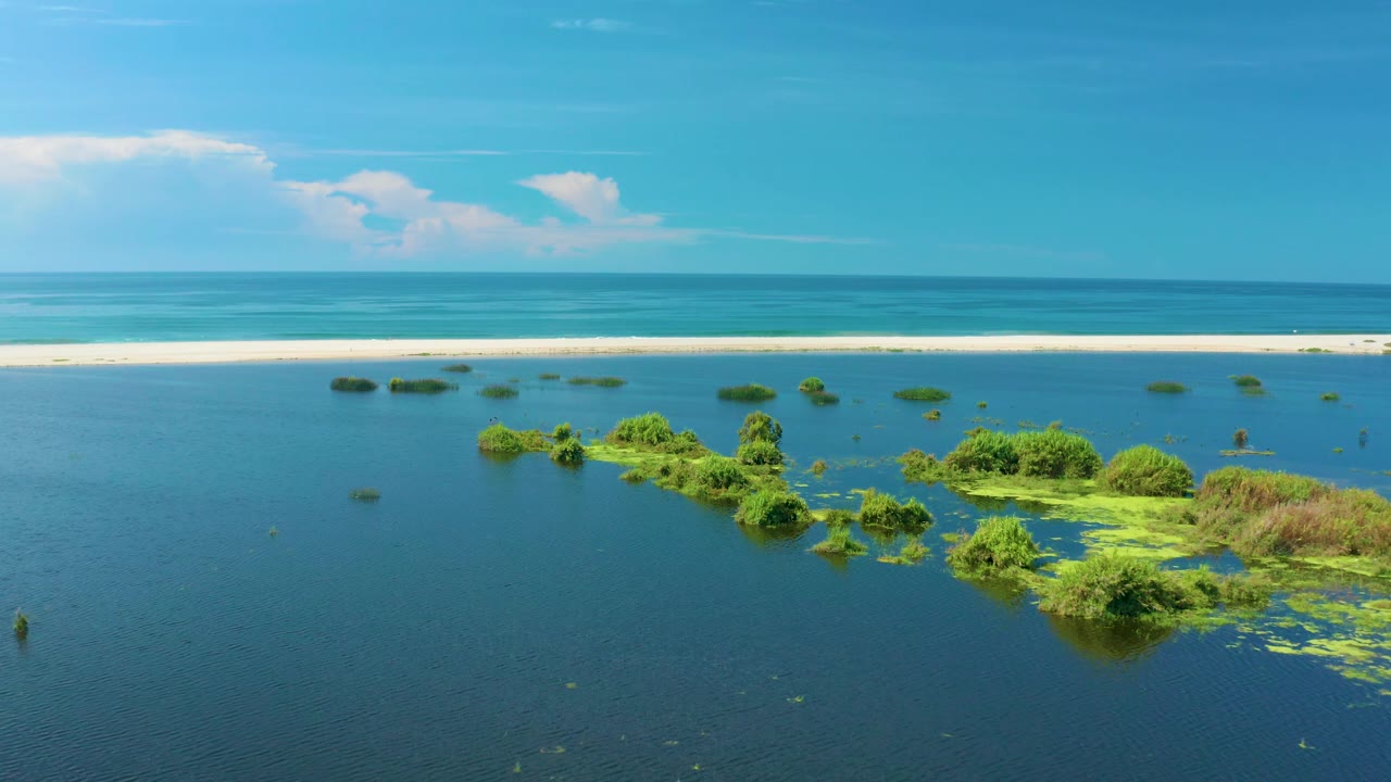Aerial coastal view in San Jose Del Cabo, Mexico, with lagoon islands lower in frame, a narrow beach and surf behind them, blue ocean, high clouds, and no visible people.