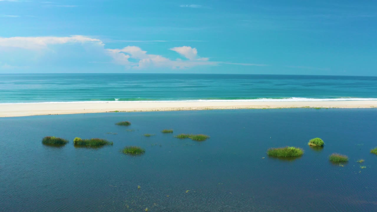 Aerial frame from San Jose Del Cabo, Mexico showing blue lagoon water with scattered grass islets, a pale sandbar, white surf, open ocean, low clouds, and no visible people.