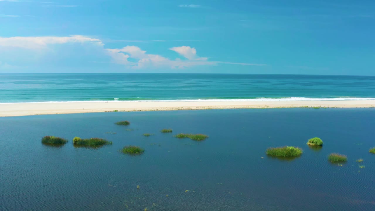 Aerial frame from San Jose Del Cabo, Mexico with clustered green grass patches across blue water, a bright sand strip, white breaking waves, ocean horizon, and no visible people.