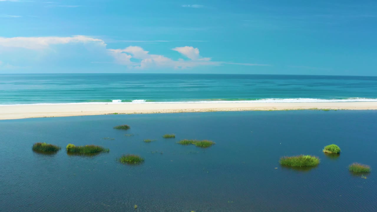 Aerial frame from San Jose Del Cabo, Mexico showing a wider spread of green islets in the lagoon, a narrow sandy beach, white surf, deep blue sea, and no visible people.