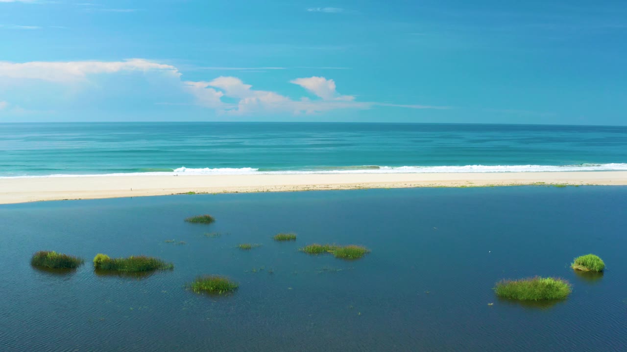 Aerial view of dark blue lagoon water, grass clumps, sandbar, and white surf.