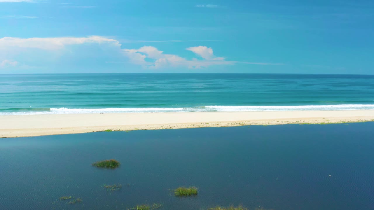 High aerial view in San Jose Del Cabo, Mexico with a broad dark lagoon foreground, narrow sandbar, rolling white surf, blue-green ocean, horizon, clouds, and no visible people.