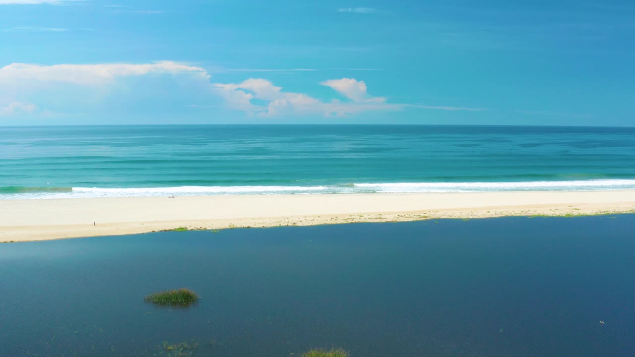 San Jose Del Cabo aerial frame with dark inlet water below, a pale sandy strip, foamy surf, calm turquoise sea, a straight horizon, scattered clouds, and no visible people.