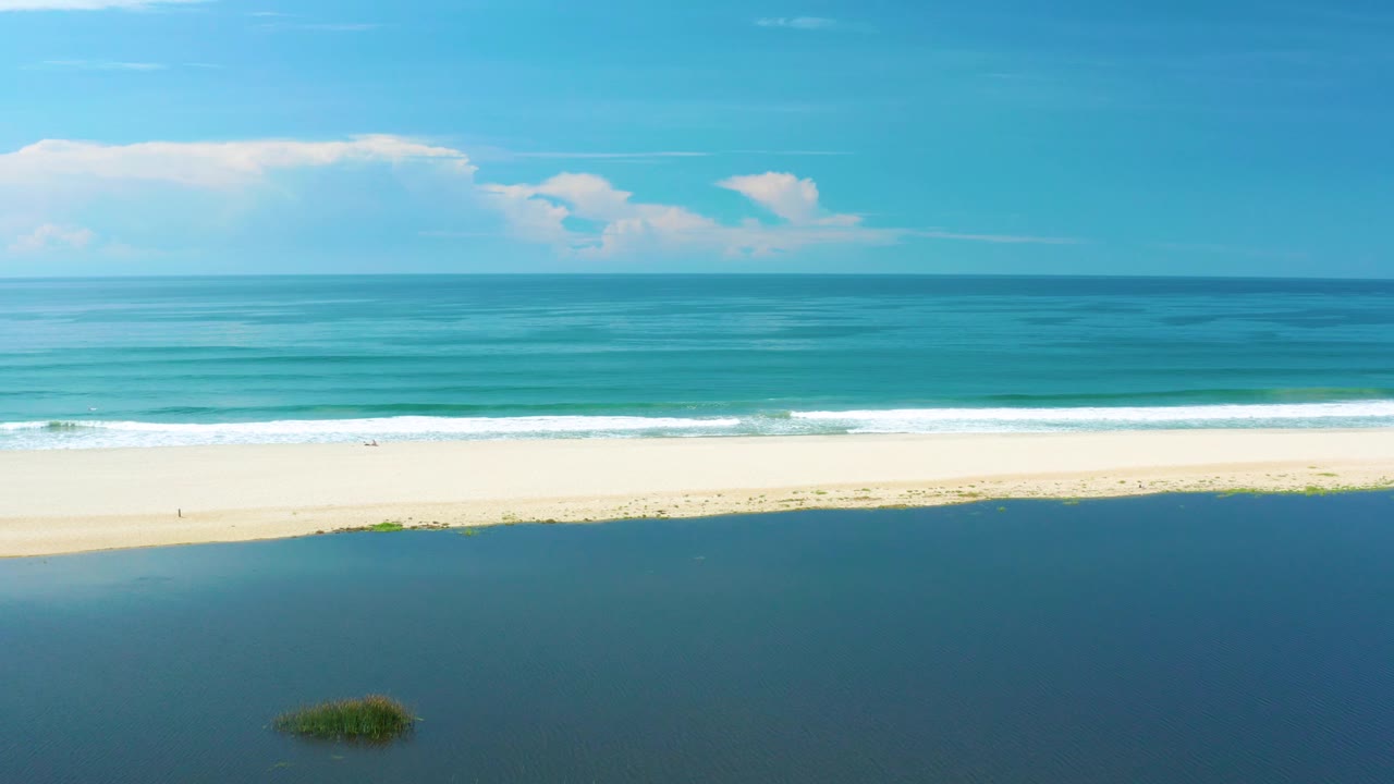 Wide aerial view over San Jose Del Cabo, Mexico with lagoon water at the bottom edge, a long sandy beach, white surf bands, blue sea, horizon, low cloud mass, and no visible people.