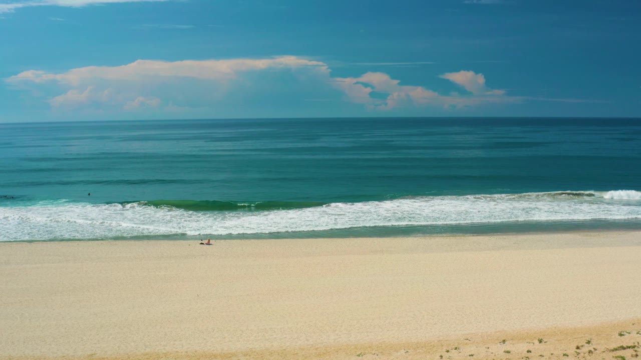 A white surf band crosses the San Jose Del Cabo beach while calm turquoise water and a clouded horizon fill the background, with no visible people.
