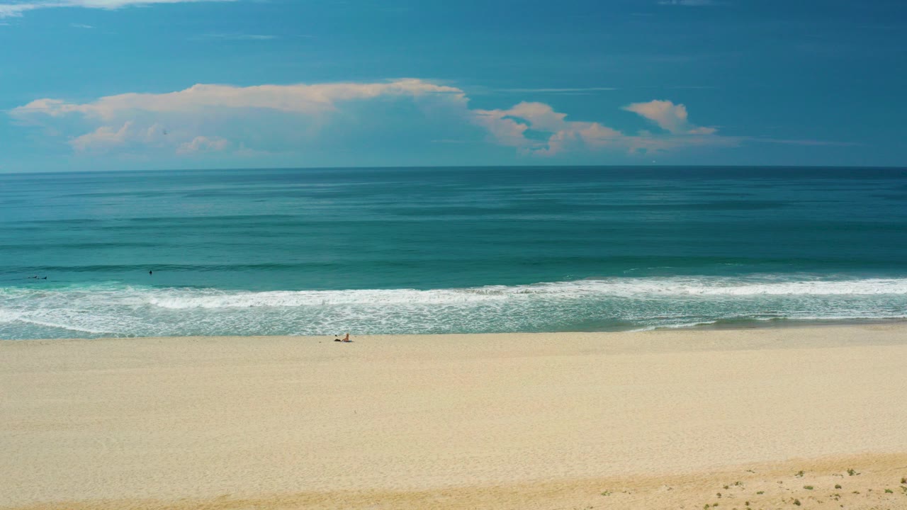 Opening frame of San Jose del Cabo beach with pale sand below a straight surf line, blue-green ocean, large white clouds, and no visible people.