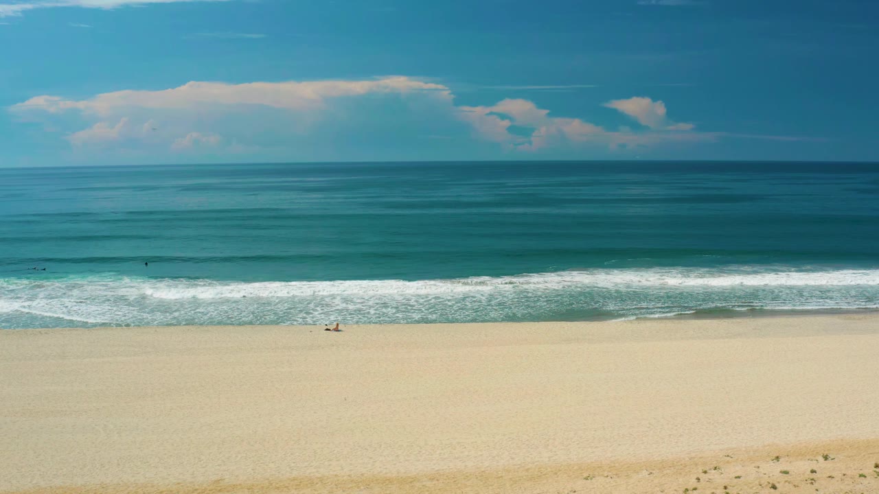San Jose del Cabo shoreline with a wide band of sand, breaking white surf across the center, open blue water, cloud bank near the horizon, and no visible people.