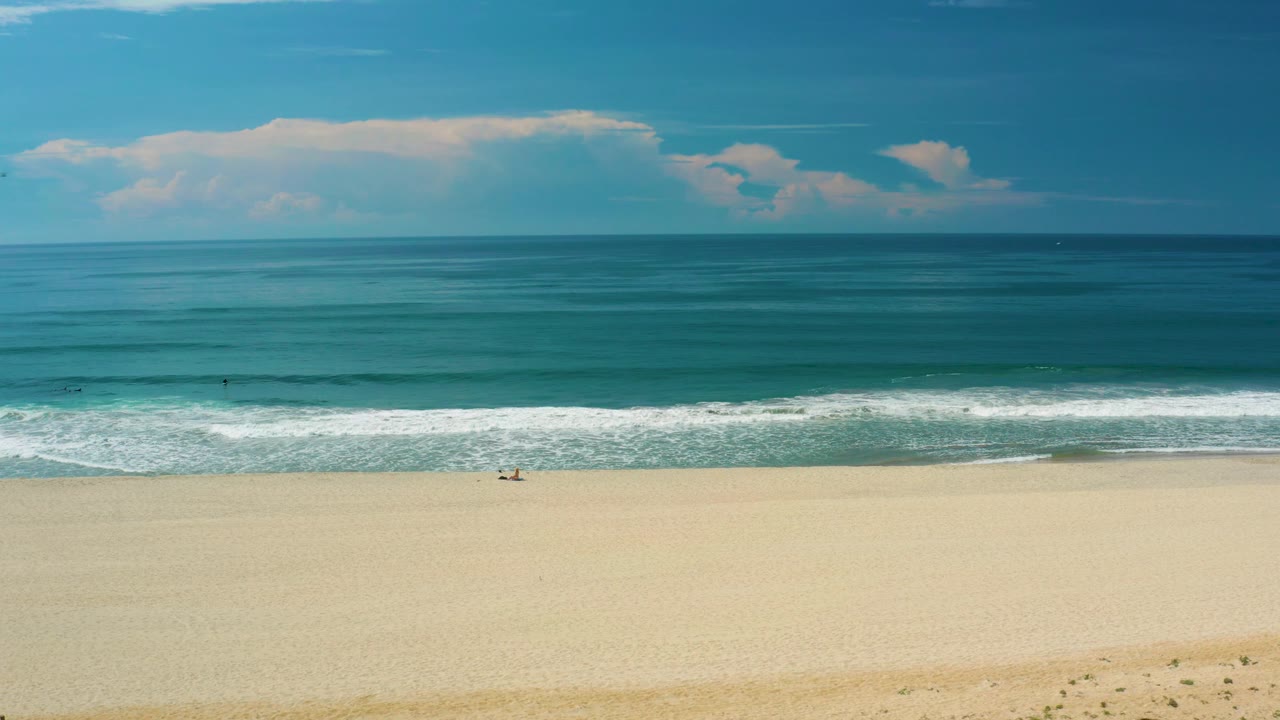 Wide San Jose del Cabo beach view with uninterrupted sand in the foreground, a bright foamy wave band, deeper blue ocean behind it, white clouds, and no visible people.