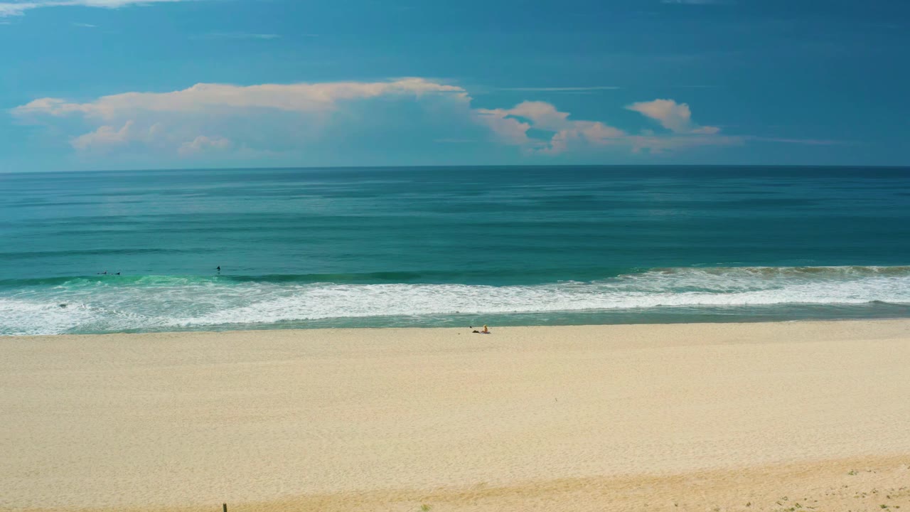 San Jose del Cabo shoreline with a smooth sand foreground, a bright band of breaking waves, open ocean, scattered cloud, and no discernible people.