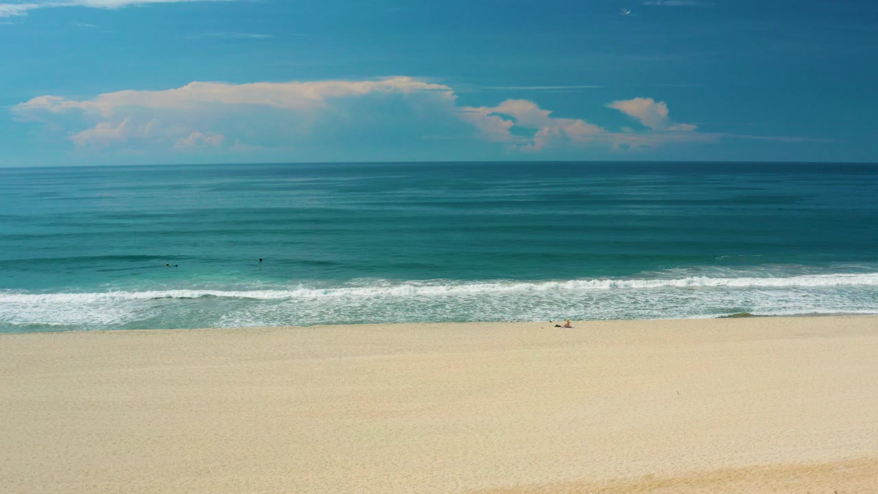 Wide view of San Jose Del Cabo beach with pale sand in front, a white surf line across the middle, teal water beyond it, a low cloud bank near the horizon, and no visible people.
