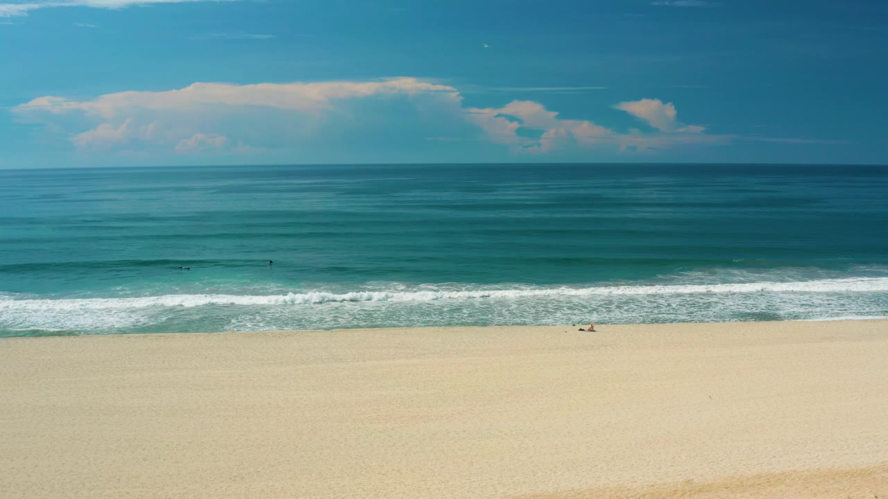 A wide San Jose Del Cabo coastal view shows layered teal water, a bright white shorebreak, pale sand across the lower frame, distant clouds, and no visible people.