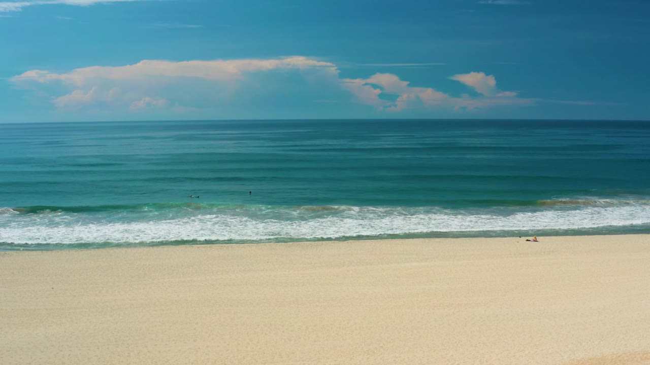 Elevated San Jose Del Cabo beach frame with turquoise water filling the center, white wave lines near shore, pale sand at the bottom, and a cloud mass above the horizon.