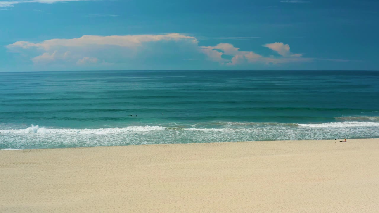 San Jose Del Cabo shoreline frame with a broad empty beach in the foreground, low white waves near shore, calm teal water, and no visible people.