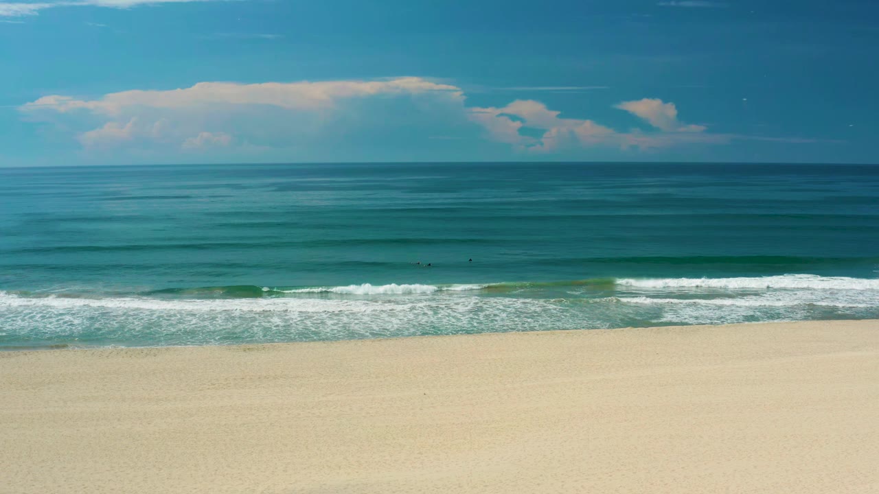 San Jose Del Cabo shoreline with a broad pale beach foreground, small white breakers near shore, blue-green water bands, and no visible people.