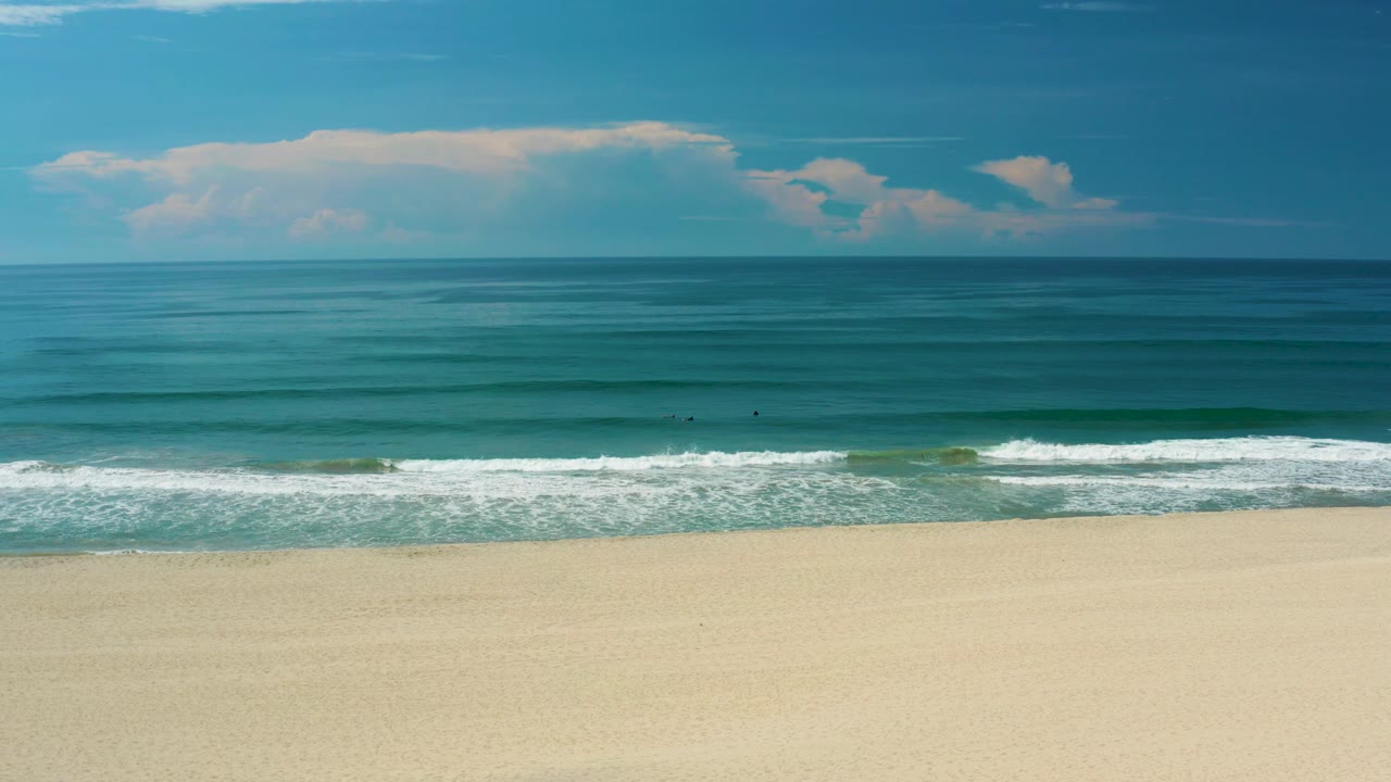 Wide San Jose Del Cabo ocean view with smooth blue water beyond the surf, a pale sandy foreground, scattered low clouds, and no visible people.