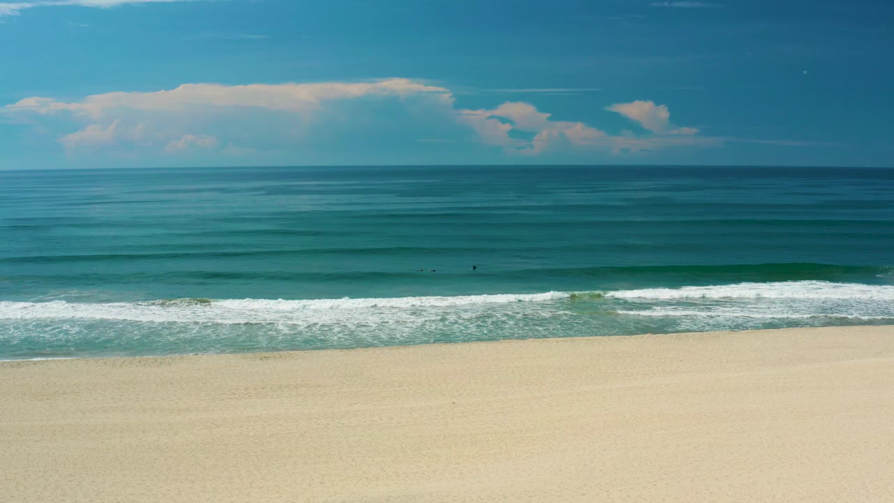 San Jose Del Cabo beach frame with white foam spread along the shore, green-blue water behind it, broad empty sand below, and no visible people.