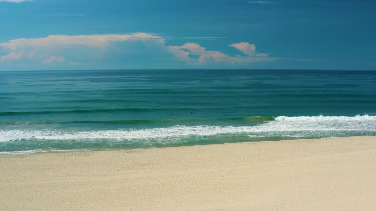 Wide view of San Jose Del Cabo beach with pale sand, turquoise water, a white surf band, distant clouds, and no visible people.