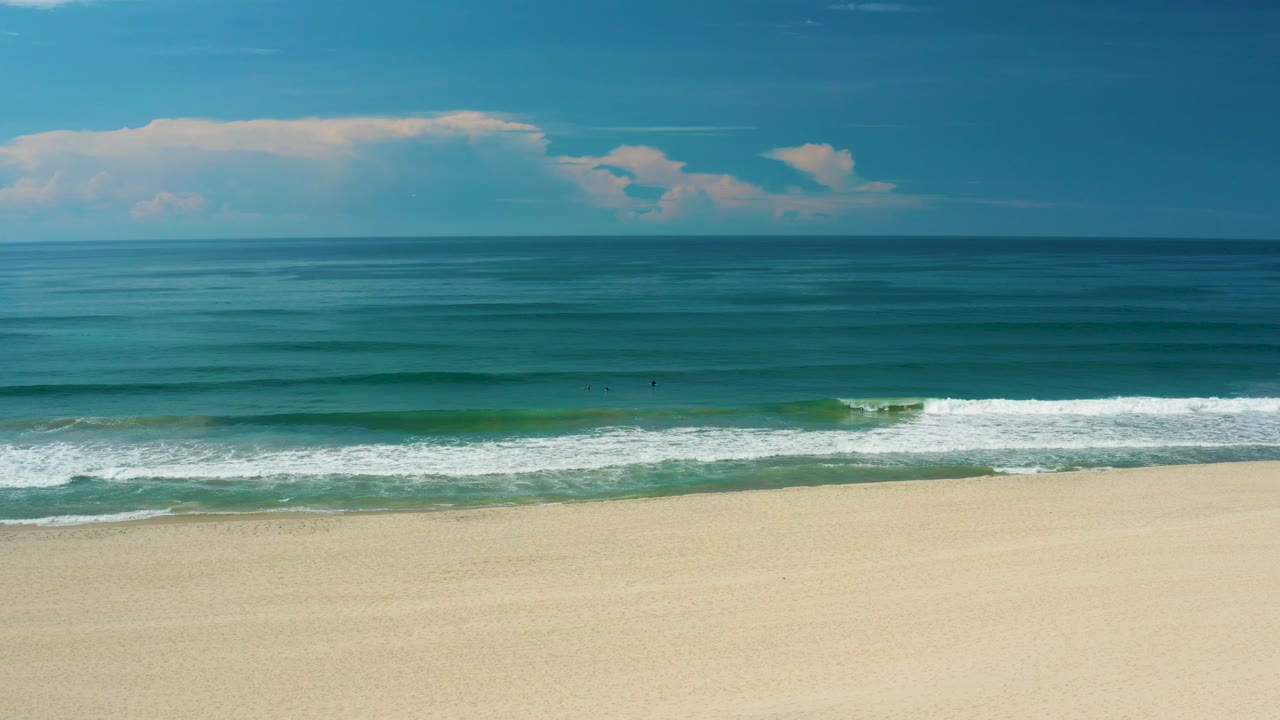 Wide San Jose Del Cabo beach frame with white foam spread near shore, darker blue water beyond, high horizon, and no visible people.