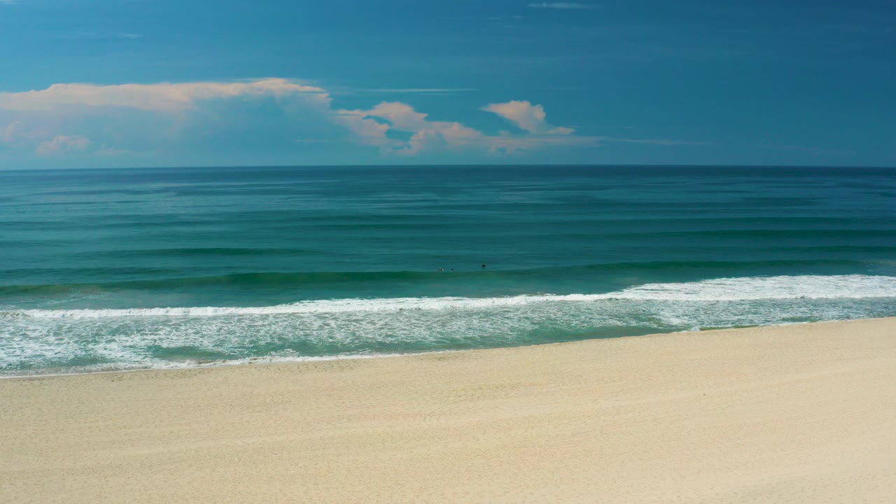 Opening wide view of a San Jose Del Cabo beach with pale sand below, a white surf line across the middle, blue ocean beyond, low clouds, and no visible people.