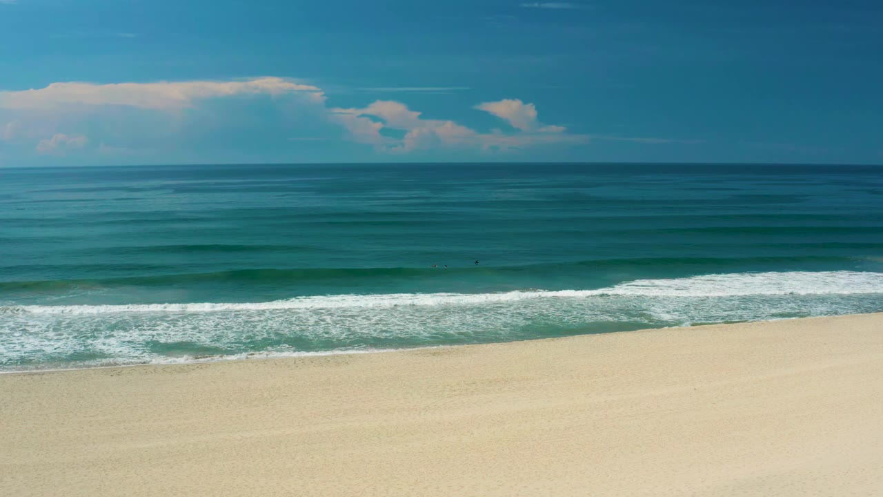 San Jose Del Cabo shoreline with smooth pale sand in the lower frame, a broad white breaking wave, layered blue water, distant clouds, and no visible people.