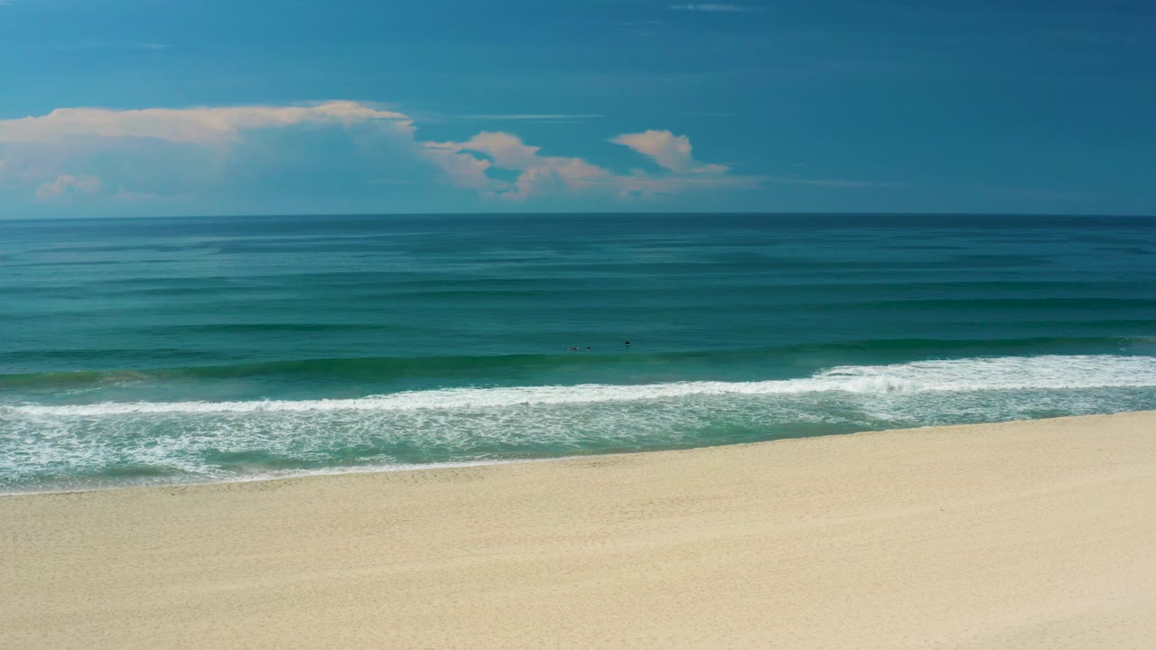 Wide San Jose Del Cabo beach frame with a slightly advanced surf line, pale sand foreground, teal water bands, a level horizon, soft clouds, and no visible people.