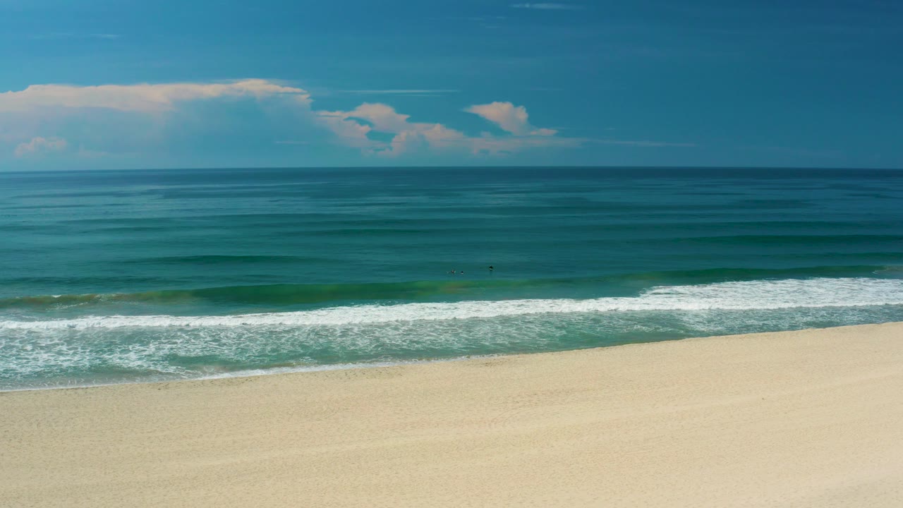 San Jose Del Cabo coast seen wide, with white foam stretching along the lower ocean edge, open sand in front, dark and light blue water bands, clouds, and no visible people.