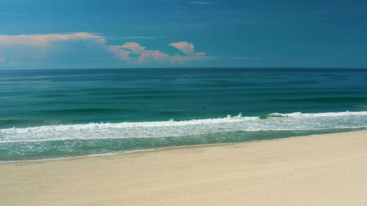 Opening frame of the San Jose del Cabo beach clip, with pale sand below white surf bands, teal ocean to the horizon, scattered clouds, and no visible people.