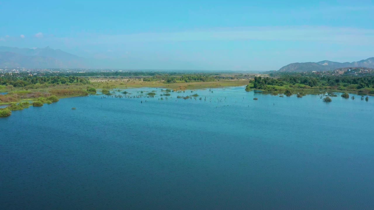 Aerial view one second in shows more open blue water in the foreground, low marsh plants across the middle, distant San Jose Del Cabo development, mountains, and no visible people.