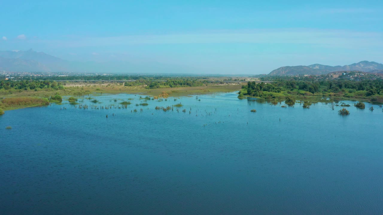 Aerial view in San Jose Del Cabo with lake water dominating the lower frame, marshy shoreline and green vegetation in the midground, hazy mountains behind, and no visible people.