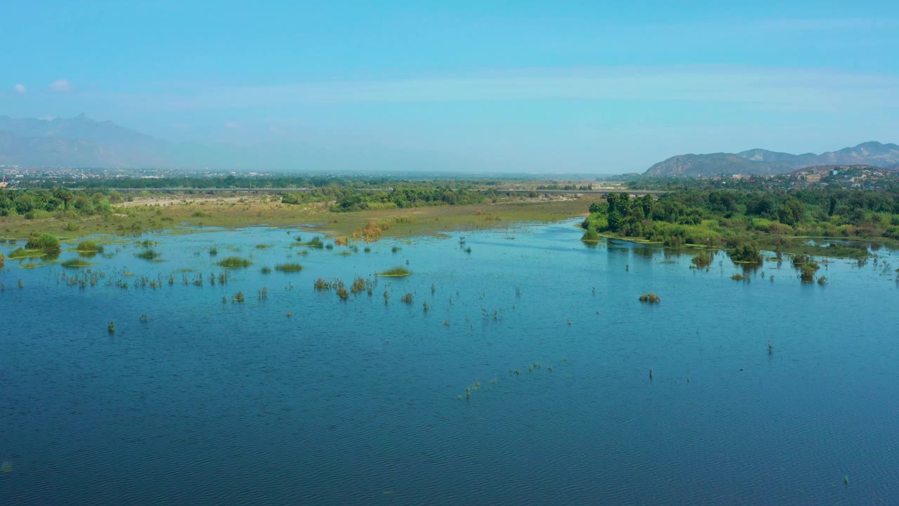 Aerial frame in San Jose Del Cabo, Mexico showing rippled blue water, scattered plant clumps, a green marshy bank, faint distant structures near the horizon, mountains, and no visible people.