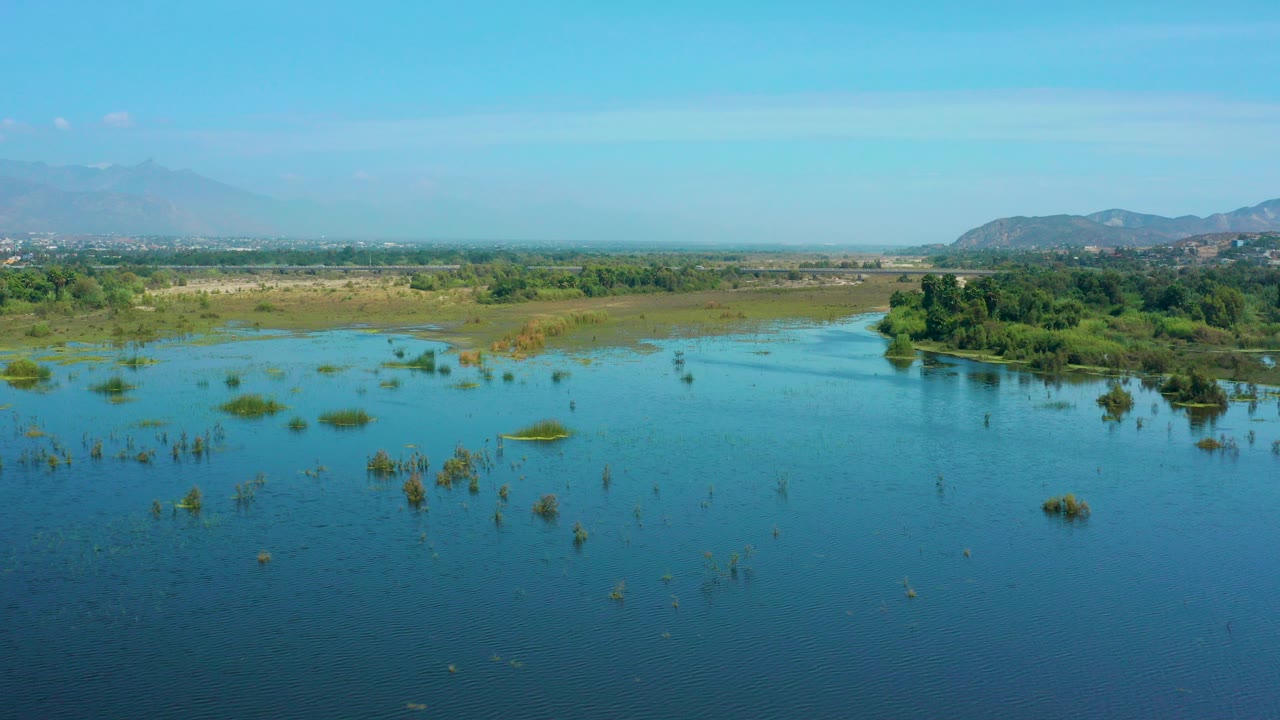Wide aerial frame over blue water in San Jose Del Cabo, Mexico, with scattered wetland bushes, a green tree island to the right of center, hazy hills in the distance, and no visible people.