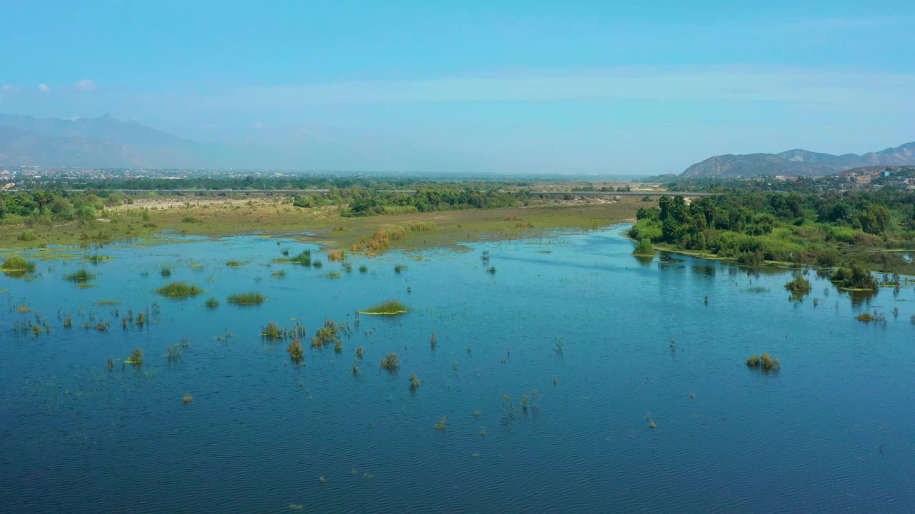 Aerial view in San Jose Del Cabo, Mexico, where blue water and small vegetation patches lead toward a tree-covered midground and faint hills under a pale sky, with no visible people.