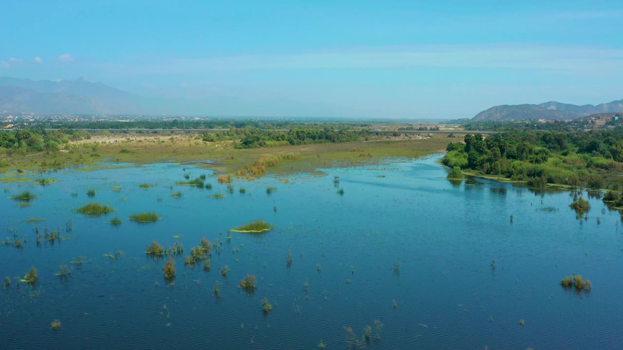 Aerial landscape of water, wetland grasses, green trees, distant hills, and bright sky.