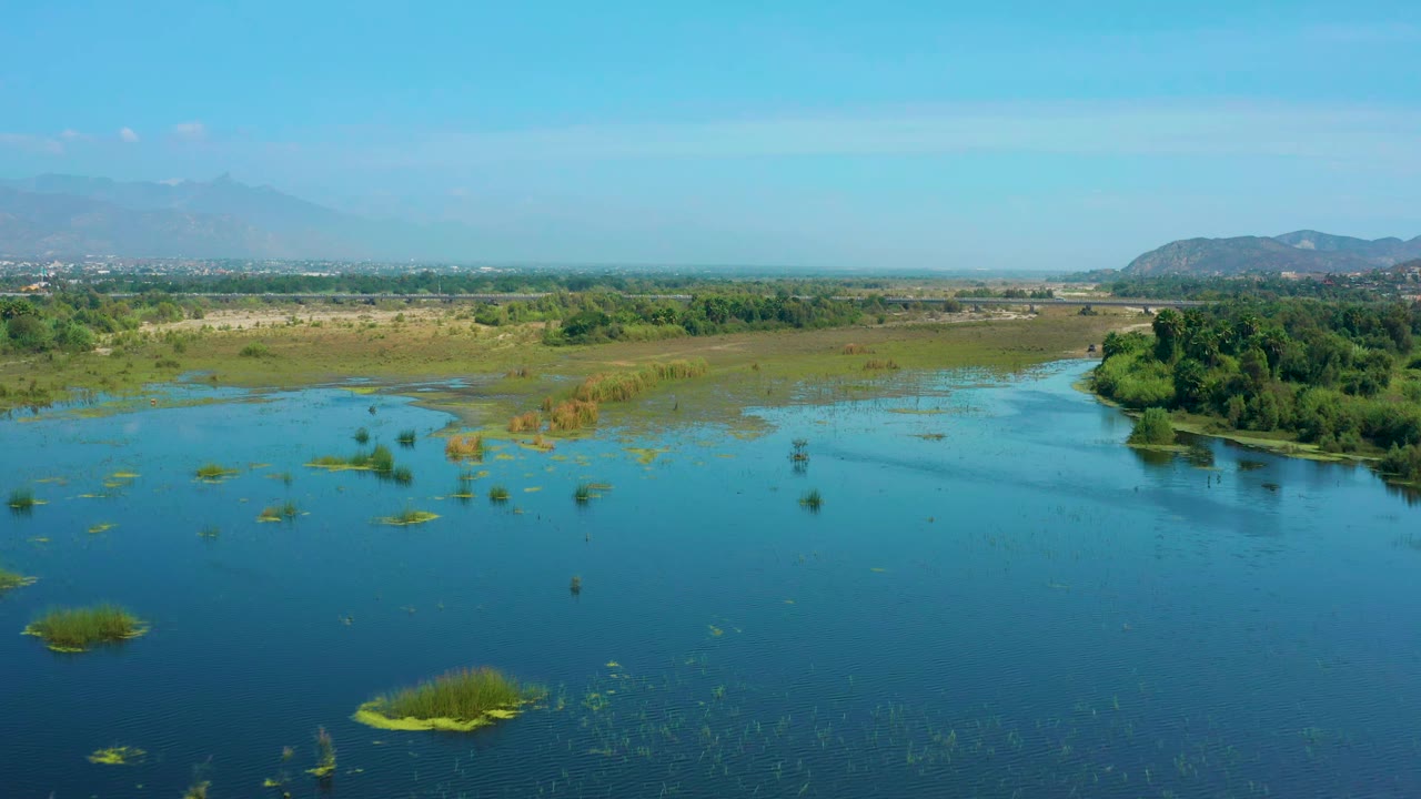 Aerial view in San Jose Del Cabo, Mexico, looking across open blue wetland water with scattered reeds, a darker green tree line to the right, hazy hills beyond, and no visible people.