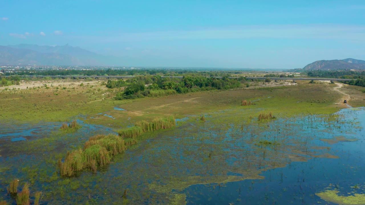 High aerial frame of San Jose Del Cabo marshlands with reflective blue water in the foreground, green vegetation bands, distant brush, hills, and no visible people.
