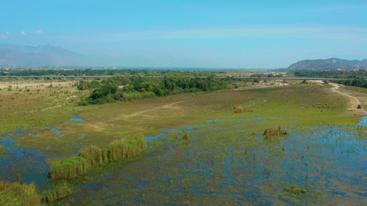 Wide aerial frame over San Jose Del Cabo wetland with blue shallow water in the foreground, tan scrubland and a dirt road beyond, hazy mountains on the horizon, and no visible people.