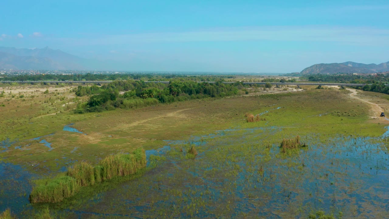 Wide San Jose Del Cabo aerial still with water channels cutting through marsh grass, scrubland across the center, a dirt track near the right edge, distant mountains, and no visible people.