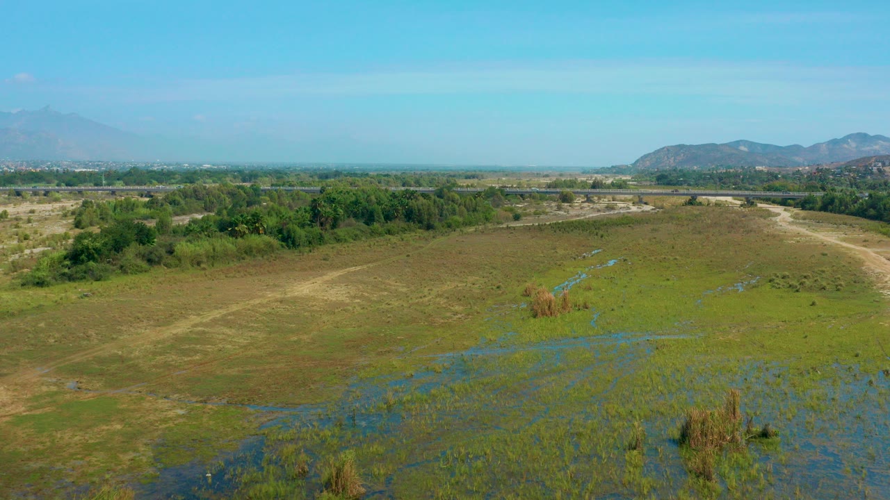 Aerial view over San Jose Del Cabo marshland with blue water channels in the foreground, a tree line and low mountains beyond, and no visible people.