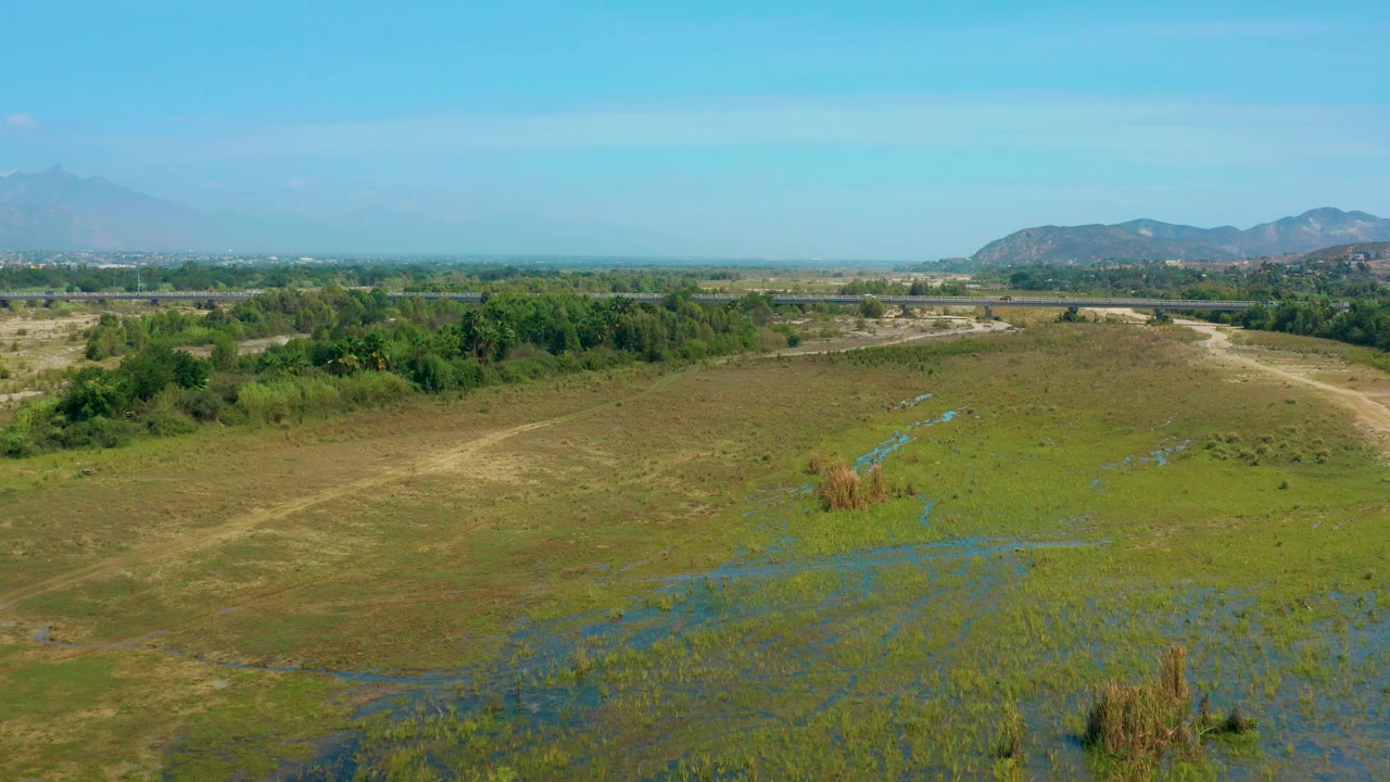 Aerial view in San Jose Del Cabo showing a diagonal dirt path through green-brown marshland, scattered blue water patches, distant hills, and no visible people.